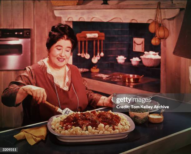 Woman prepares to serve pasta shells with tomato sauce from a large casserole dish while standing behind a counter in a kitchen.