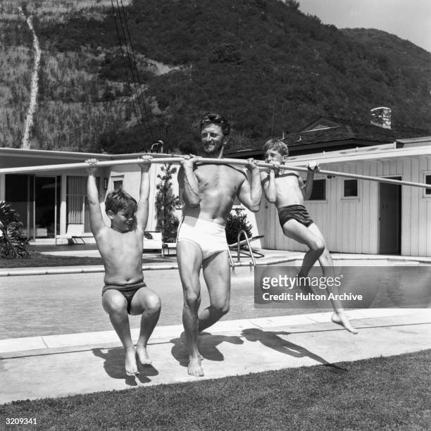 Full-length image of American actor Kirk Douglas using a pole to lift his sons, Joel and Michael, on the deck of a swimming pool.