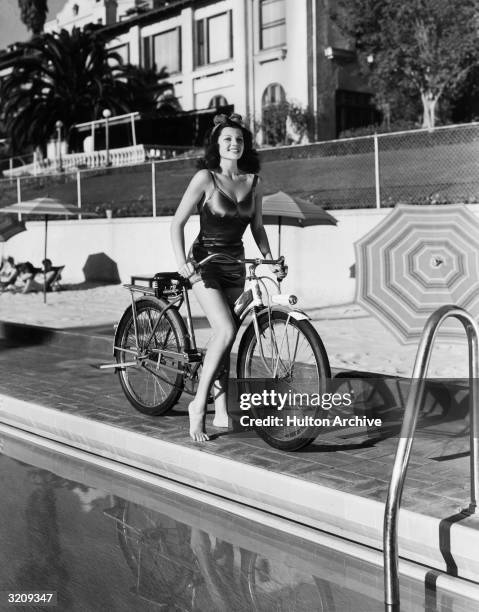 American actor Rita Hayworth poses on a bicycle in a swimsuit by the side of a swimming pool at the Beverly Hills Hotel, California.