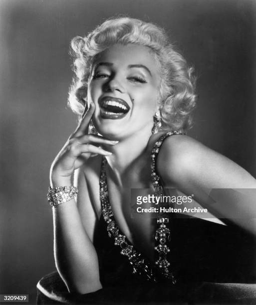Half-length portrait of American actor Marilyn Monroe laughing, her hand raised to her cheek, wearing a low cut dress trimmed in jewels.