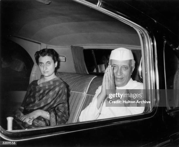 Indian prime minister Pandit Nehru clasping his hands in salute while sitting with his daughter, Indira Gandhi, in the backseat of a car.