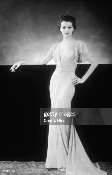 Portrait of a woman leaning on a wall while modelling a long chiffon dress with a plunging V-neck, ruffled cap sleeves, and a fishtail train.