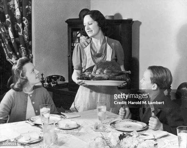 Mother, wearing an apron, presents a whole cooked turkey on a platter to her daughter and son at their dining room table.