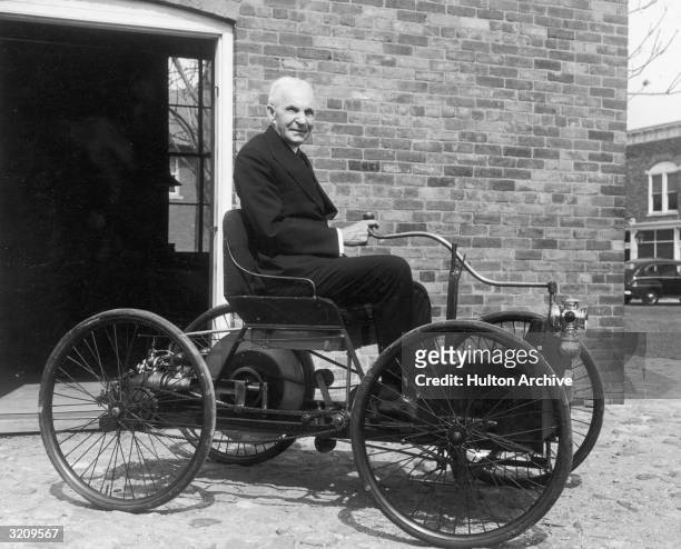 Portrait of American inventor and industrialist Henry Ford sitting in the first automobile he built, a 'quadricycle', in front of his former...