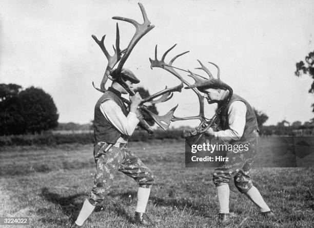 Two men, wearing traditional costumes, face off while holding deer antlers over their shoulders outdoors in a field, Abbots Bromley, Staffordshire,...
