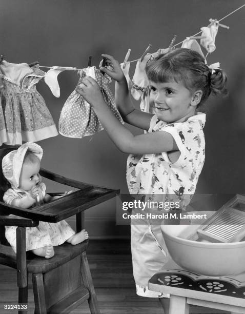 Young girl smiles as she hangs doll clothes on a clothesline, with her baby doll in a high chair beside her. There is a washboard and basin on a...