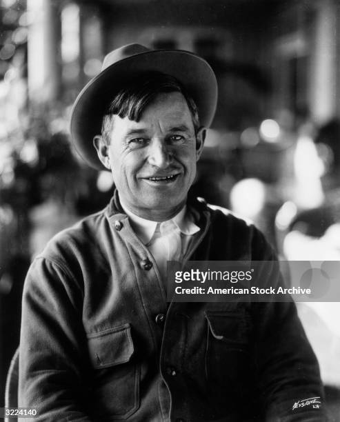 Portrait of American entertainer Will Rogers sitting and smiling. He wears a hat and an overshirt.
