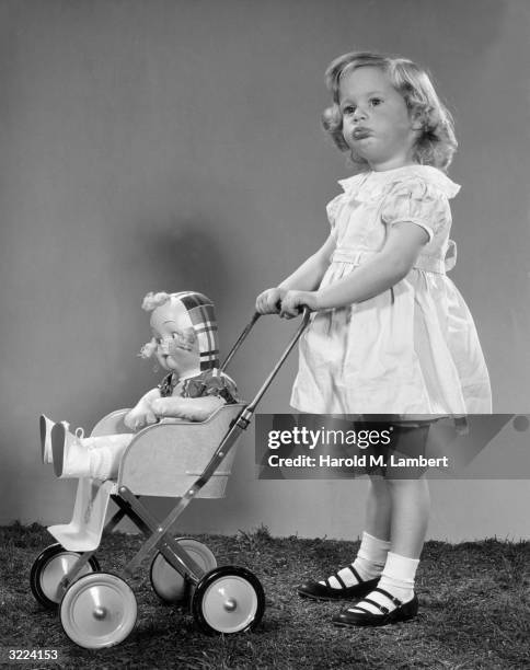 Full-length studio image of a young girl standing on grass while pushing a female doll in a baby carriage.