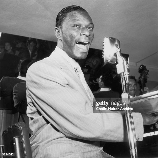American pop singer and pianist Nat 'King' Cole plays piano and sings into a microphone. An unidentified bassist plays behind him.