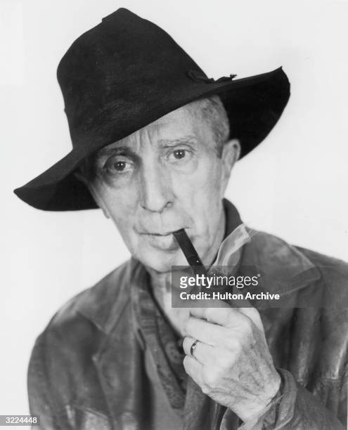 Studio headshot portrait of American painter Norman Rockwell smoking a pipe while wearing a floppy felt hat.