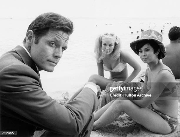 American actor Jack Lord sitting on a beach next to two women in bikinis, in a still from the television show, 'Hawaii Five-O'.