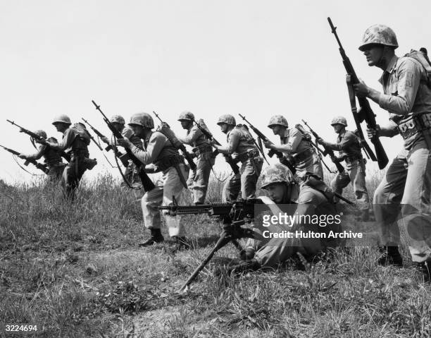 Line of U.S. Marines advances, most brandishing M-14 rifles and one kneeling behind an M-60 machine gun.