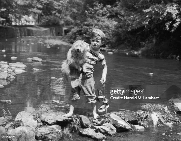 Boy in rolled-up jeans crosses a stream barefoot while carrying his hightop sneakers and a sheepdog.