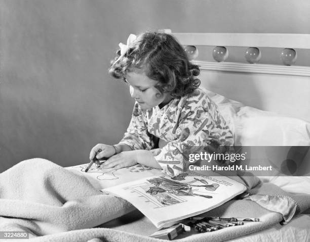 Young girl sits in bed, leaning forward as she colors with crayons in a coloring book. The box of crayons lays open on her blanket.