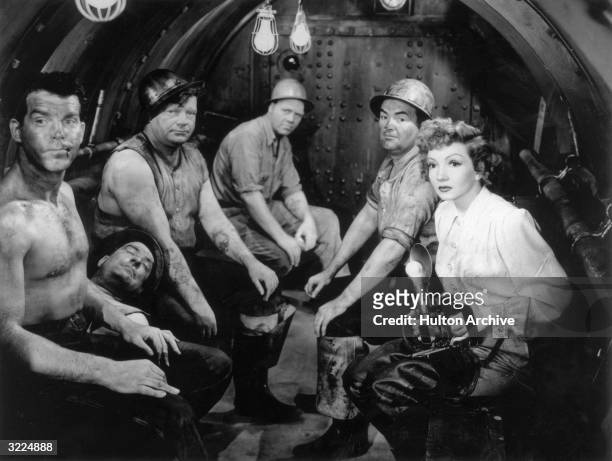 French-born actor Claudette Colbert holds a camera as she sits with a group of dirty men in a tunnel, including American actor Fred MacMurray , in a...