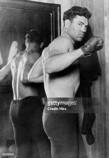 American heavyweight boxing champion Jack Dempsey , wearing boxing gloves and tights, poses in fighting stance next to a mirror.