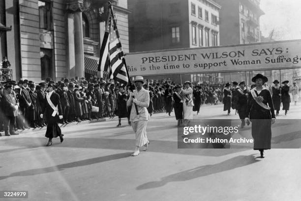 Full-length image of suffragettes marching with a banner declaring, 'PRESIDENT WILSON SAYS: This is the time to support Woman Suffrage,' New York...