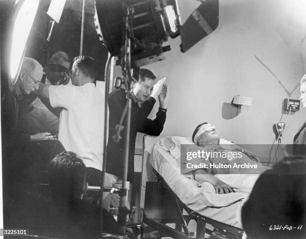 American film director Roger Corman lines up a shot as Welsh-born actor Ray Milland lays in a hospital bed with a bandage over his eyes, on the set...