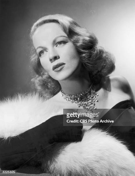 Studio portrait of actor Maria Riva wearing a black evening gown and a fur stole, looking up.
