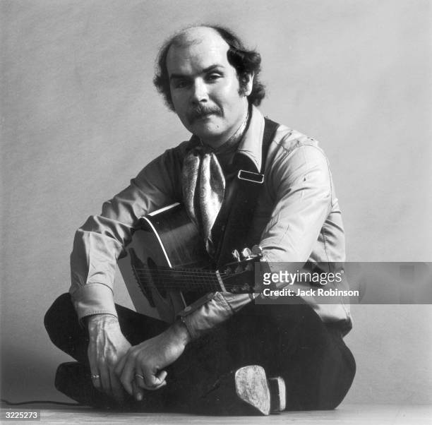 Full-length studio portrait of American folk singer and songwriter Tom Paxton sitting cross-legged with his acoustic guitar.