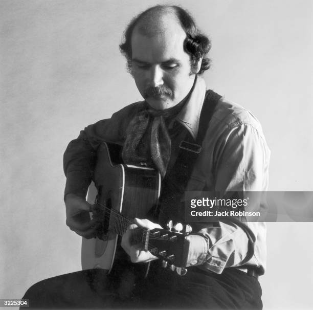 Studio portrait of American folk singer and songwriter Tom Paxton sitting and playing his acoustic guitar.