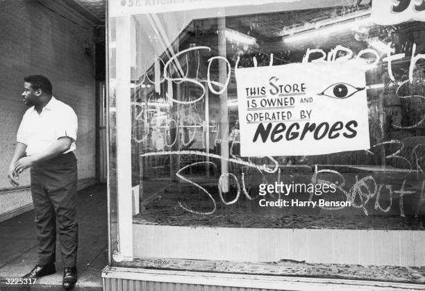 An African-American storekeeper stands outside his shop, which has a sign reading, 'This store is owned and operated by Negroes,' during the race...