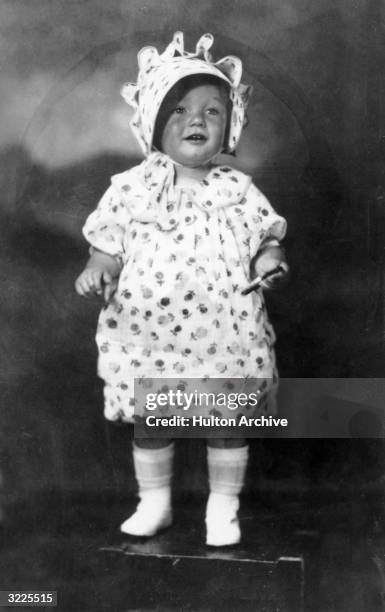 Full-length studio portrait of American actor Marilyn Monroe at the age of two, standing on a box in a floral print dress and matching bonnet.