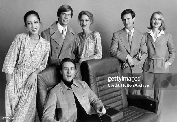 Studio portrait of American fashion designer Halston sitting in front of models wearing his clothes, which are made from coordinated fabrics.