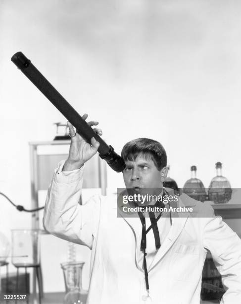 American actor, comedian and director Jerry Lewis, wearing a lab coat in a laboratory, looks through the wrong end of a telescope in a promotional...