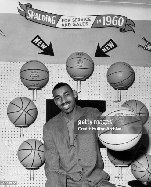 American basketball player Wilt Chamberlain wearing a business suit, smiling and seated at the Spalding basketball display at the opening of a...