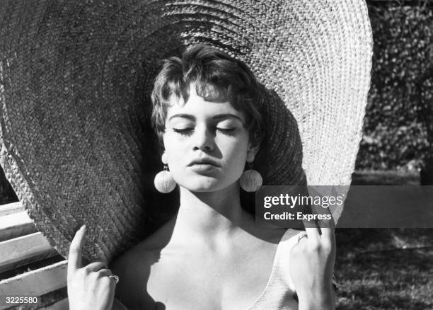 Headshot of French actor Brigitte Bardot closing her eyes and holding the rim of her straw hat while basking in the sun outdoors. Bardot has brunette...