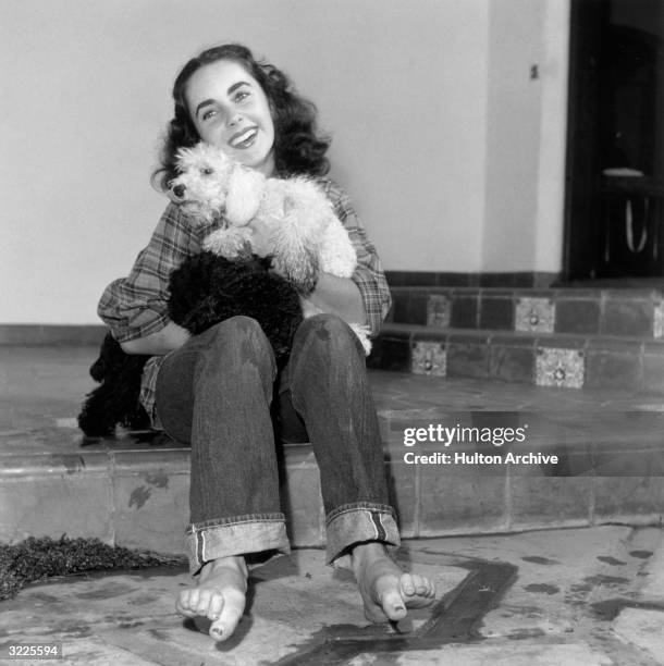 British-born actor Elizabeth Taylor, wearing a plaid shirt and jeans rolled up at the cuff with bare feet, holding a poodle and smiling.