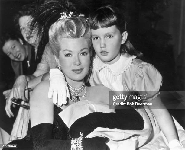 American actor Lana Turner, wearing a period costume with a feather headdress, sits with her daughter, Cheryl Crane, on the set of director Curtis...