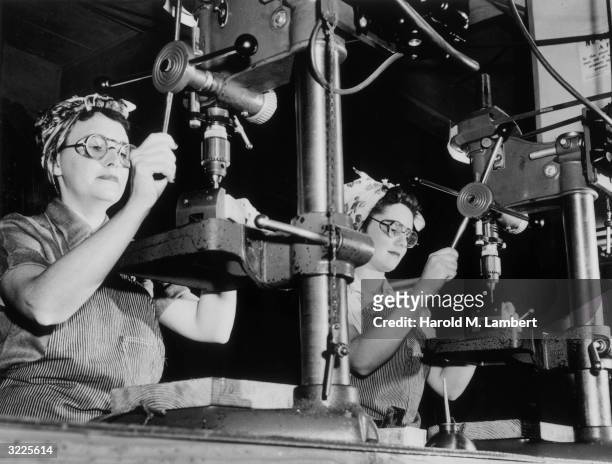 Two female machinists use drill presses while working side-by-side in a homefront factory during World War II. Both women wear goggles, headscarves,...