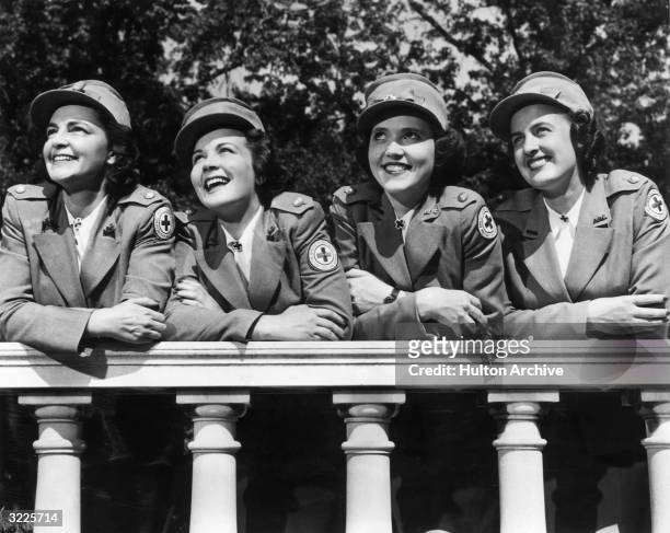 Four female members of the American Red Cross stationed in England smile as they lean over a wall, wearing their uniforms and caps, London, England....