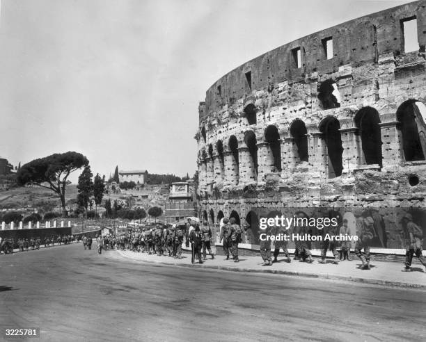 United States troops walk down a curved street past the Coliseum in Rome, Italy. A Roman gentleman walks in the opposite direction, using a cane. The...