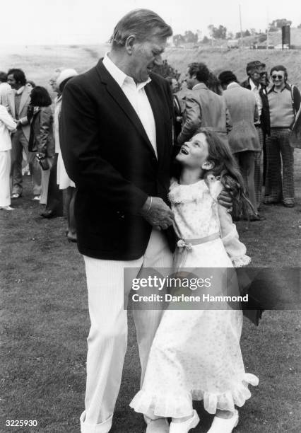 Full-length image of American actor John Wayne with his arms around his daughter, Aissa, who is smiling up at him. Behind Wayne and his daughter are...