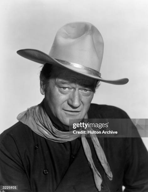 Headshot portrait of American actor John Wayne dressed as a cowboy with a bandana around his neck and a white cowboy hat on his head.