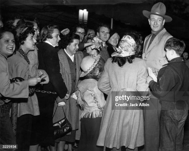 American actor John Wayne dressed in a suit and a cowboy hat while surrounded by a crowd of fans the night he immortalised his handprints and...