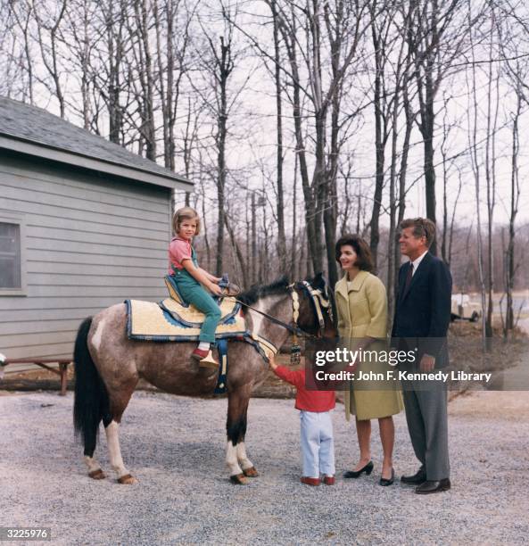 President John F Kennedy stands outdoors with his wife, First Lady Jacqueline Bouvier Kennedy , as their daughter, Caroline, sits atop a pony and...