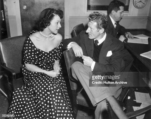 American singer Kay Starr, in a polka-dotted dress, sits talking to American actor Danny Kaye while an unidentified man reads at a desk behind them....