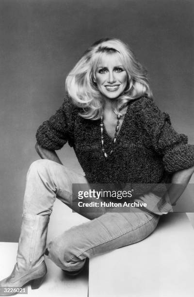 Studio portrait of American actor Suzanne Somers sitting with her hands on her hips. She is wearing corduroy jeans tucked into cowboy boots, a...
