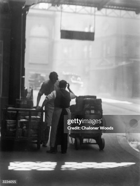 Two porters having a conversation while leaning on their trolleys which are full of parcels.