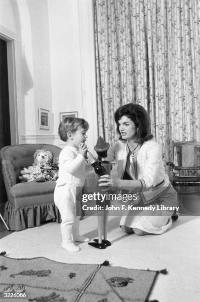 American First Lady Jacqueline Bouvier Kennedy kneels on the floor with her son, John F. Kennedy, Jr. , playing with a wooden toy figure, inside the...