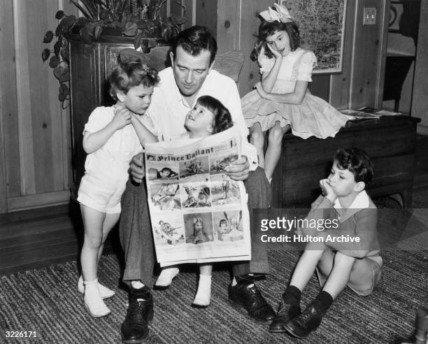 American actor John Wayne sitting on a chair and reading a Prince Valiant comic with his four children, Hollywood, California. From left: Patrick,...