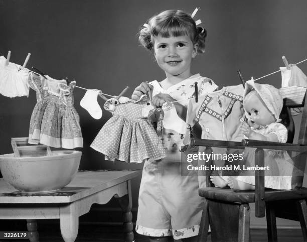 Studio portrait of a young girl with pigtails standing behind a clothesline, with doll clothes hung on the line. There is a bowl with a small...