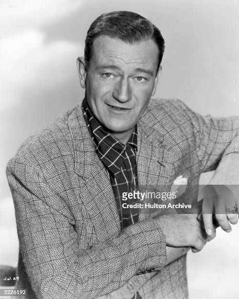Promotional portrait of American actor John Wayne , posing in a checkered sports jacket with his elbows resting on a wooden branch.