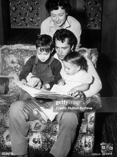 Married American actors John Derek and Pati Behrs read a book with their son, Russell, and their daughter, Sean. Derek sits on a couch and reads the...