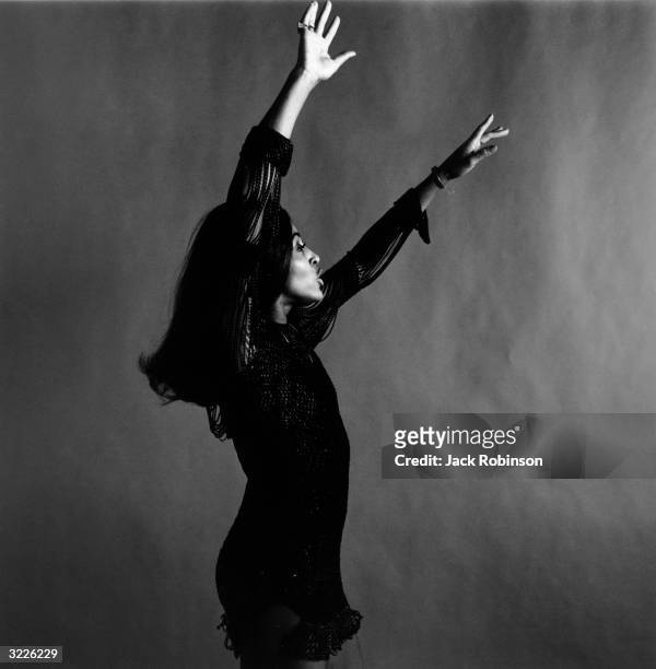 Studio portrait of rock singer Tina Turner, wearing a dark crocheted mini-dress, dancing in profile with her arms over her head while singing, New...