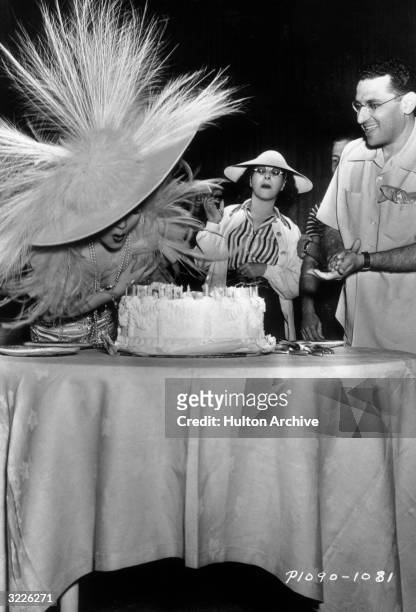 French-born actor Claudette Colbert blows out the candles on her birthday cake as American film director George Cukor, American costume designer...